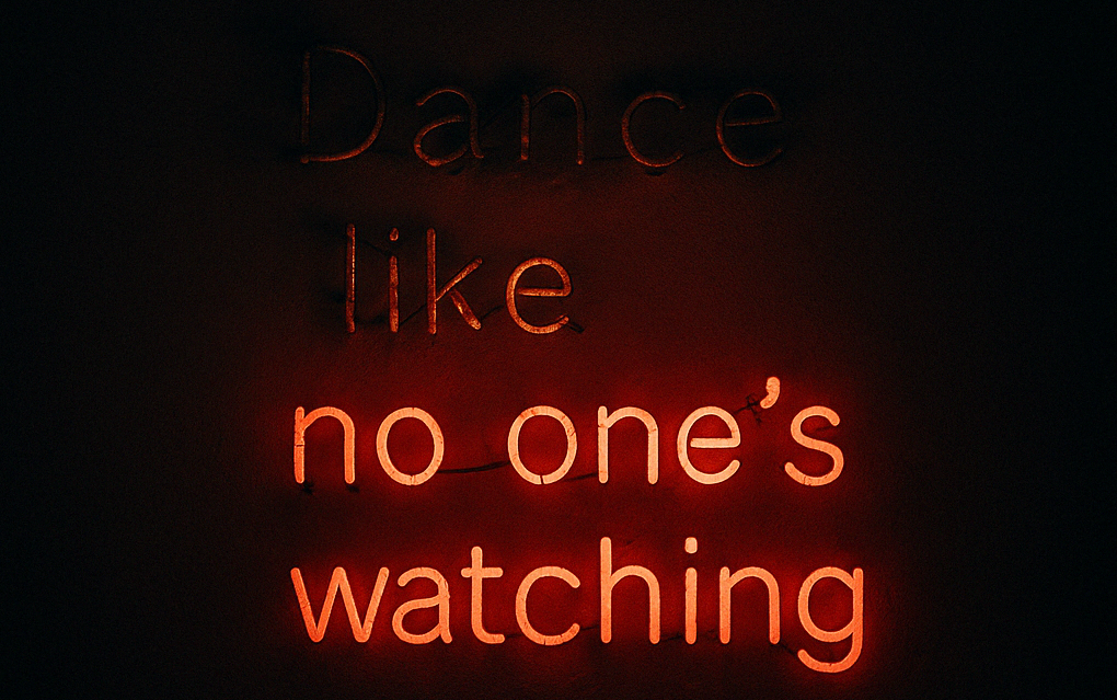 A neon sign on a dark wall that says “Dance like no one’s watching.” The words "dance like" are barely glowing. The rest glows softly in red, casting a faint light.