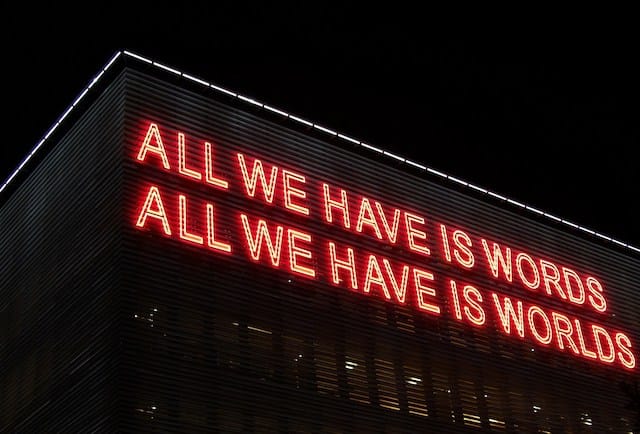 Red neon sign reading 'ALL WE HAVE IS WORDS ALL WE HAVE IS WORLDS' on a dark building at night.