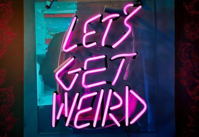 Bright pink neon ‘LET’S GET WEIRD’ sign mounted on dark wall.